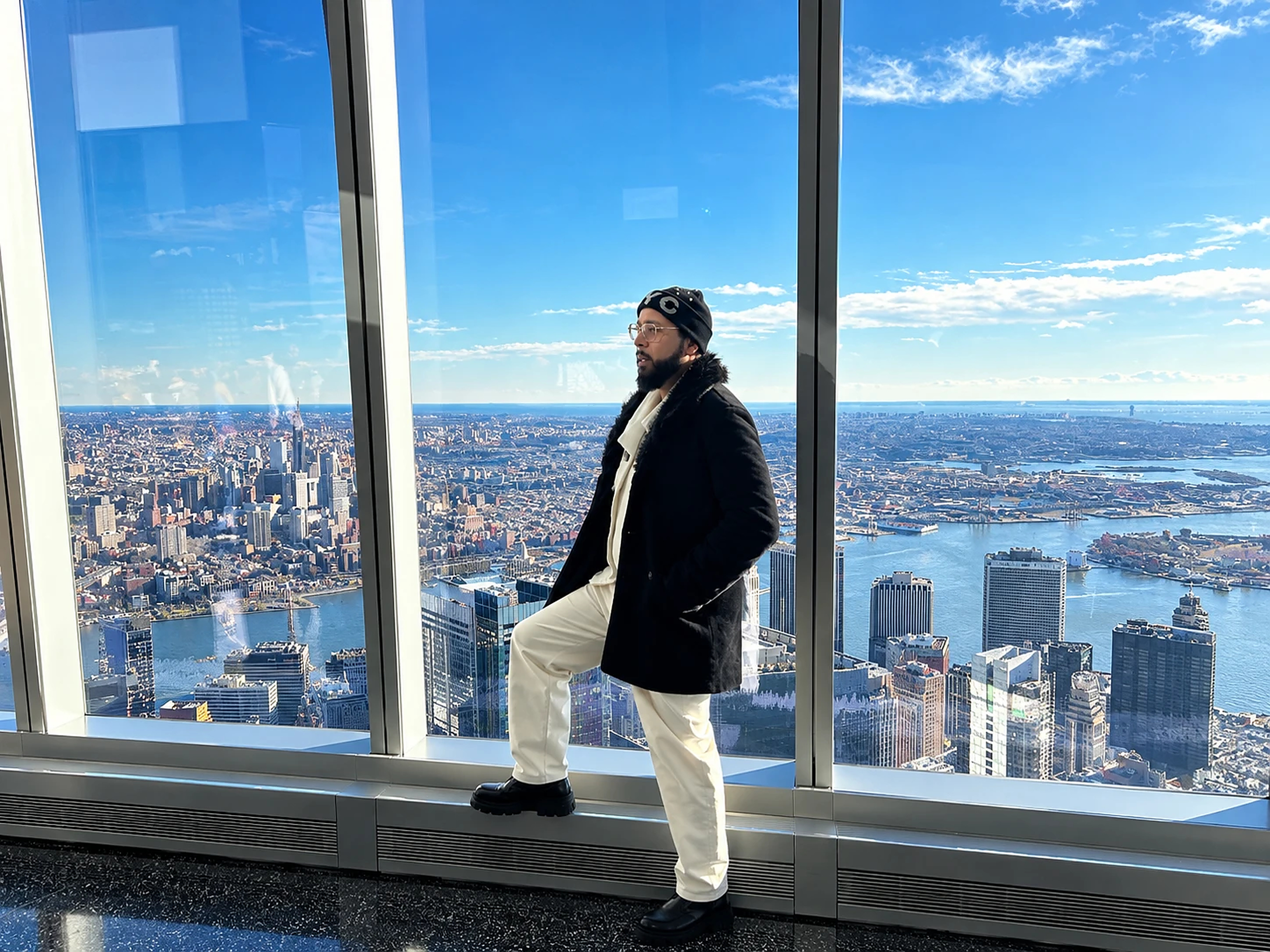 Carlos Romero in a black coat and cream trousers at a skyline window, New York